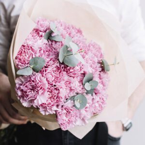 A bouquet of pink carnations with eucalyptus leaves, wrapped in beige paper.