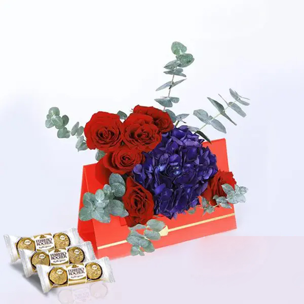 01 (7) A red card with red roses, blue hydrangeas, and eucalyptus leaves, accompanied by a box of chocolates on the table below.