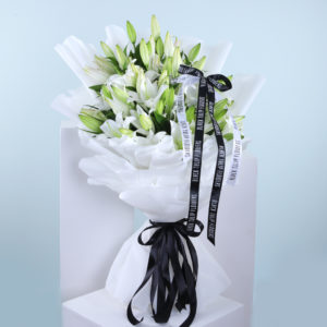 A large bouquet of pristine white lilies, elegantly wrapped in white tissue paper and tied with a black ribbon.