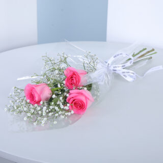 Small bouquet of 3 pink roses with baby's breath and greenery in a transparent wrap tied with white ribbon.