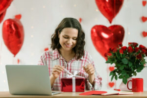 A young woman with a joyful expression is seated at a table with a laptop unwrapping a gift