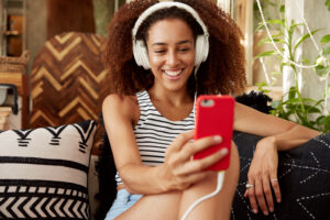beautiful-young-woman-has-afro-hairstyle-makes-video-call-via-smart-phone-headphones-speaks-with-friend-online-while-sits-comfortable-sofa-with-cushions