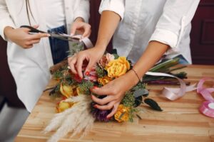 Two people, likely florists, working together to create a colorful bouquet of flowers on a wooden table. One person is cutting a ribbon while the other arranges the flowers.
