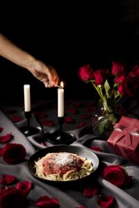 A romantic Valentine's Day dinner scene with a plate of spaghetti covered in red sauce and grated cheese, lit candles, a bouquet of red roses, and rose petals scattered on the table