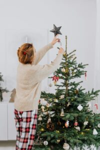 a person decorating A beautifully decorated Christmas tree with warm, golden lights. The tree is adorned with ornaments and stands in a wicker basket. Surrounding the tree are wrapped gifts in various colors and patterns, a wooden rocking horse, and a small table with a Christmas decoration. The background is a textured, light gray wall.