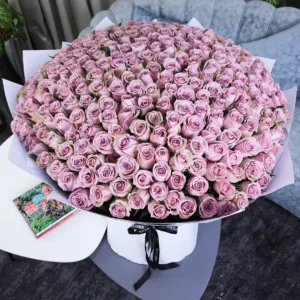 A massive bouquet of lavender or mauve roses, wrapped in white paper, with a small gift and a grey sofa in the background.