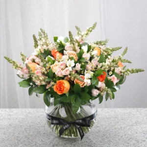 The arrangement features a variety of flowers in shades of peach, orange, pink, and white. Prominent are orange roses, light pink roses, white flowers that appear to be stocks or snapdragons, and smaller white and pink blossoms.