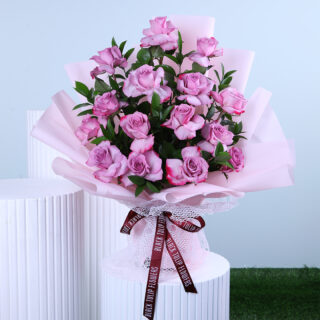 A bouquet of roses and greenery, hand-tied and wrapped in light pink tied with maroon ribbon displayed on a white pedestal.