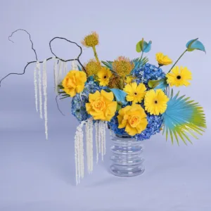 A vibrant floral arrangement in a clear, fluted glass vase against a light purple background. The arrangement features bright yellow roses and gerbera daisies, accented with blue hydrangeas and unique, spiky yellow flowers.