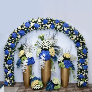 floral arch, as you suggested. The arch is composed of blue and white flowers and greenery, arranged in a semi-circular shape. It's set against a light-colored wall, creating a visually appealing contrast.