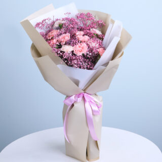 A bouquet of light pink carnations and pink baby's breath wrapped in brown kraft paper with layers of white tissue and tied with a light pink satin ribbon.