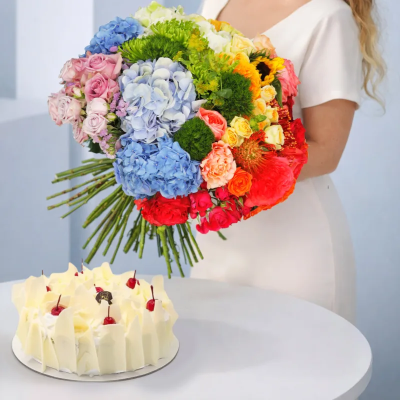mixed-bloom-white-forest-cake A gift box arrangement featuring a vibrant mixed flower bouquet with assorted fresh blooms placed neatly beside a white forest cake.