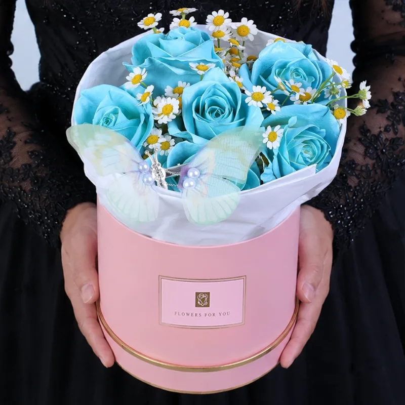 blue-rose-butterfly-arrangement A decorative floral arrangement featuring blue roses, delicate white blooms, and a butterfly accent, elegantly arranged in a pink round box.