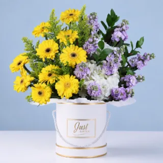 Round box flower arrangement with cheerful yellow gerberas, soft lavender blooms, white hydrangeas, and fresh ruscus leaves for a premium floral gift.