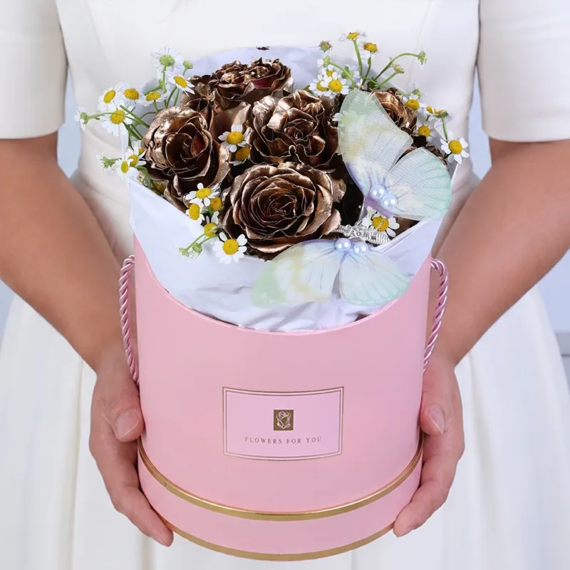 golden-butterfly-rose-round-box_2026_01 Premium box featuring gold-painted roses, white tanacetum flowers, and a butterfly placed among the blooms for a luxurious gifting experience.