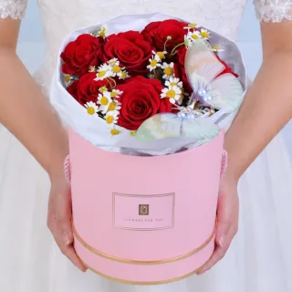 Handcrafted pink round box featuring seven red roses and delicate white tanacetum flowers with a decorative butterfly placed among the blooms, creating a charming and romantic floral arrangement.