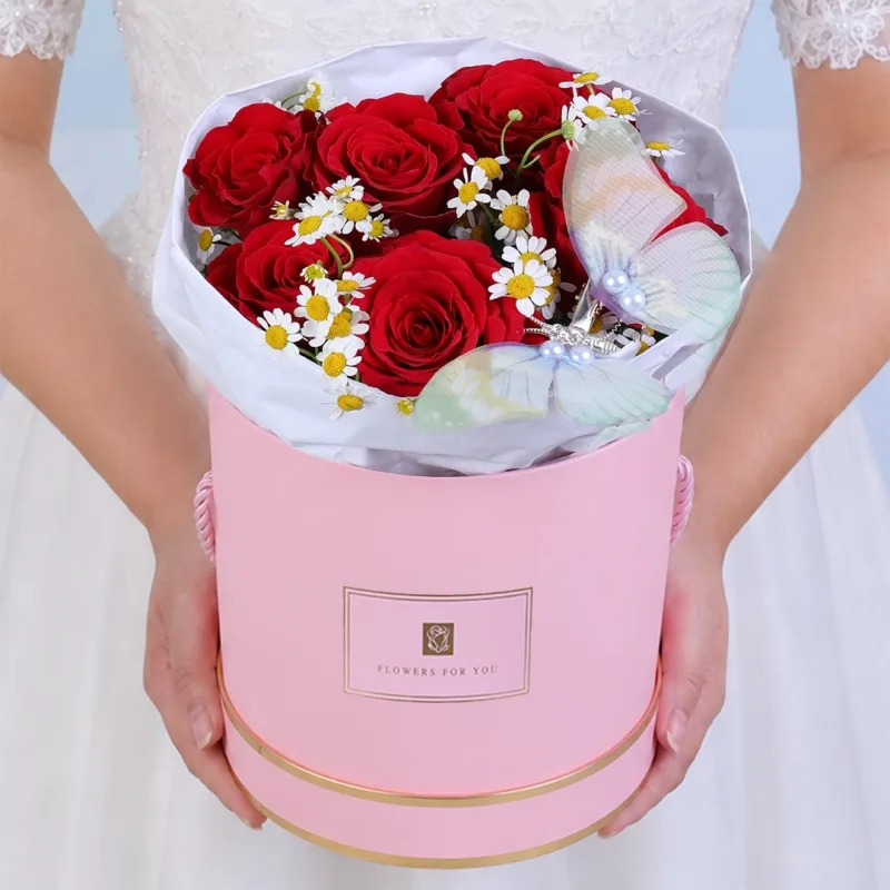 rose-blooms-and-butterfly-charm_2026_01 Handcrafted pink round box featuring seven red roses and delicate white tanacetum flowers with a decorative butterfly placed among the blooms, creating a charming and romantic floral arrangement.