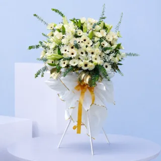 Sacred White Sympathy Stand featuring white roses, white gerberas, white lilies, gypsophila, eucalyptus baby blue, and gold-painted monstera leaves arranged on a white steel sympathy flower stand.