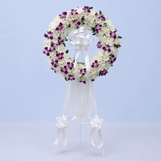 White & Purple Orchid Tribute Stand featuring white chrysanthemums, purple dendrobium orchids, and fresh single ruscus arranged in a round sympathy wreath on a white steel stand.