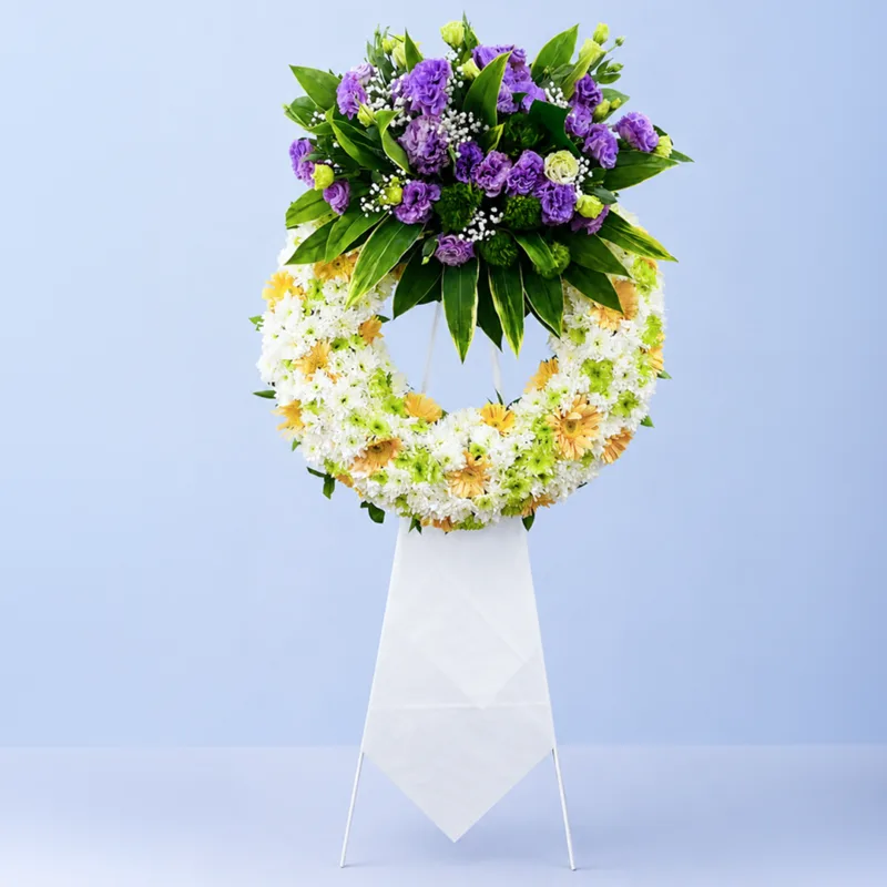 white-purple-tribute-wreath-stand_2026_01 White & Purple Tribute Wreath Stand featuring white chrysanthemums, purple lisianthus, peach gerberas, green chrysanthemums, gypsophila, and fresh greenery arranged on a white steel sympathy wreath stand.