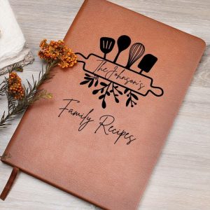 a personalized, light brown, leather-bound recipe book resting diagonally on a light gray wooden surface.