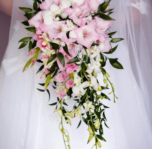 a bridal bouquet held against a white dress. The bouquet features a mix of pink and white flowers, including alstroemeria and orchids, with green leaves and delicate white embellishments.